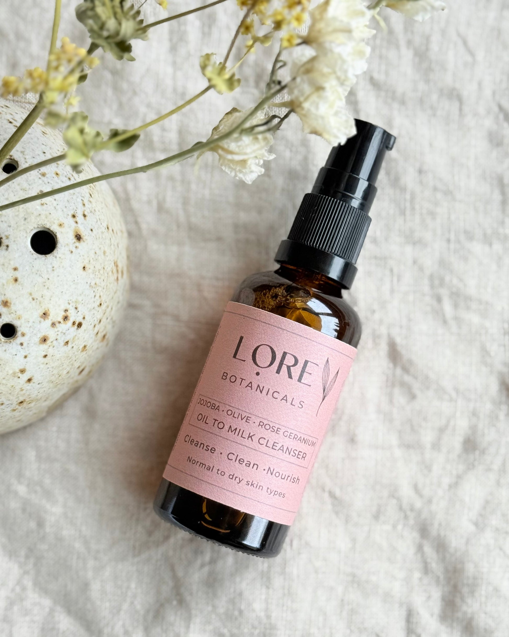 A bottle of Lore Botanicals Oil To Milk Cleanser with a spray pump, placed on a fabric surface with a floral design, alongside a ceramic dish and dried flowers.