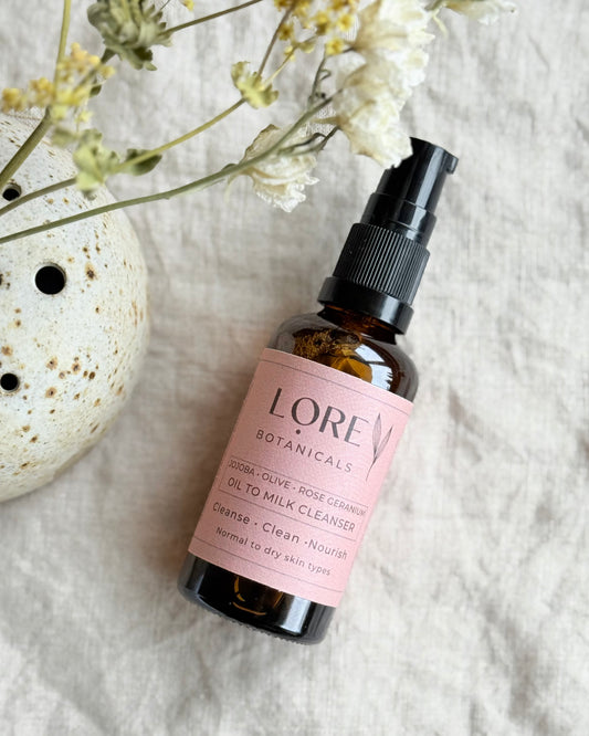 A bottle of Lore Botanicals Oil To Milk Cleanser with a spray pump, placed on a fabric surface with a floral design, alongside a ceramic dish and dried flowers.